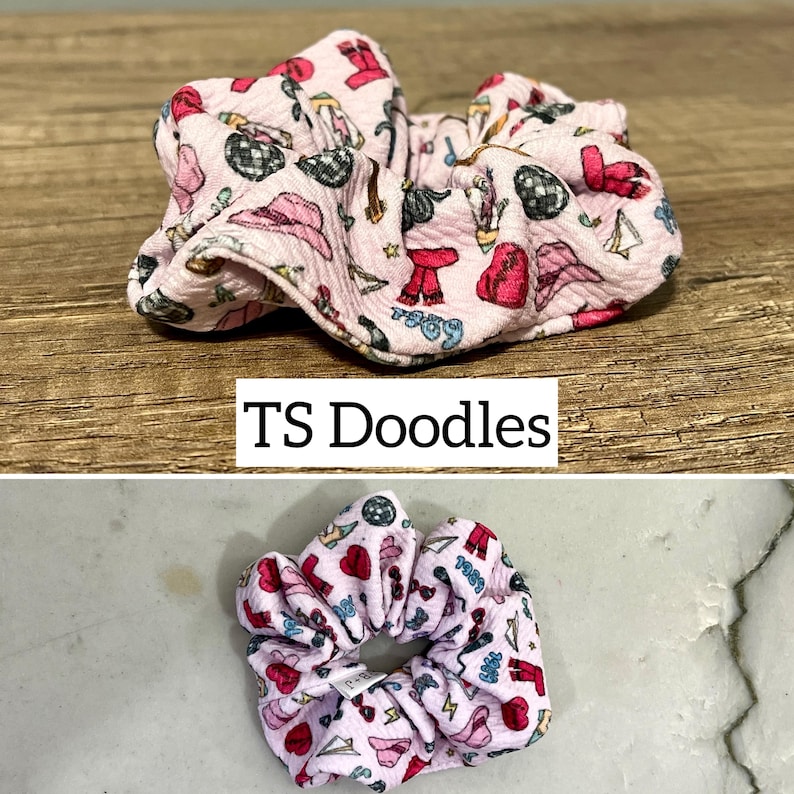 May include: Pink fabric scrunchie with a pattern of red hearts, stars, and other small designs. The scrunchie is made of a soft, textured fabric and is tied in a knot.
