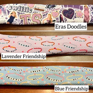 May include: Three fabric headbands with different patterns. The top headband is purple and white with a variety of images and text, including "Swiftie" and "Eras Doodles". The middle headband is lavender with a repeating pattern of hearts and the text "Swiftie" and "Karma". The bottom headband is blue with a repeating pattern of hearts and the text "Swiftie" and "Lover".