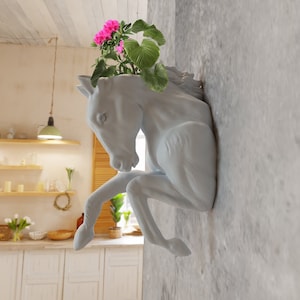 Può includere: Un vaso da fiori in ceramica bianca a forma di testa di cavallo con fiori rosa che crescono dalla parte superiore. Il vaso è montato su una parete grigia.