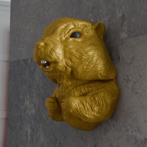 Könnte beinhalten: Eine goldfarbene Tierskulptur eines Bibers mit offenem Maul, das seine Zähne zeigt. Die Skulptur ist an einer grauen Wand befestigt.