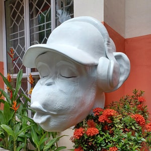 May include: White painted sculpture of a monkey wearing a white baseball cap and headphones. The monkey has its eyes closed and its mouth is slightly open. The sculpture is set against a backdrop of red and white walls and green plants.