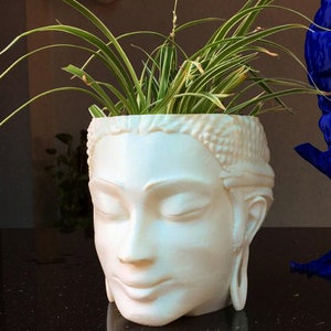 Puede incluir: Maceta de cerámica blanca con forma de cabeza humana con los ojos cerrados y una expresión serena. Una planta verde crece en la parte superior de la maceta.