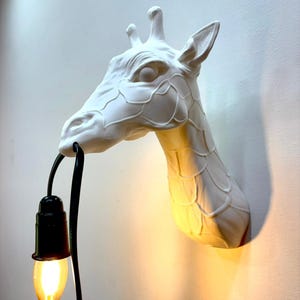 May include: A white giraffe head wall lamp with a black cord and a lit, teardrop-shaped lightbulb. The giraffe head is mounted on a white wall, casting a warm glow.