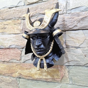 Puede incluir: Un casco de samurái negro con detalles dorados, montado en una pared de piedra texturizada. El casco presenta un rostro detallado, cuernos dorados y un detalle dorado en forma de cuerda alrededor del cuello. La pared está compuesta por piedras de varios tonos marrones, beige y grises.