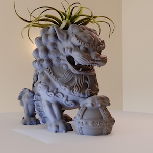 Puede incluir: Una estatua de león de piedra gris con detalles intrincados, sosteniendo un orbe redondo en sus patas. El león está decorado con una planta verde frondosa que crece de su cabeza.