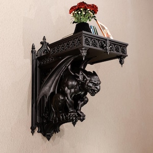 May include: A black wall-mounted shelf featuring a detailed gargoyle figure as the support bracket. The shelf has an ornate, pointed arch design along the edges. A small bouquet of red roses and a thin book are placed on the flat top surface.