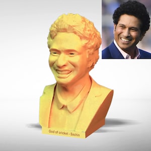May include: A yellow and orange 3D printed bust of a man with a smiling face. The bust is on a small base with the text "God of cricket - Sachin".