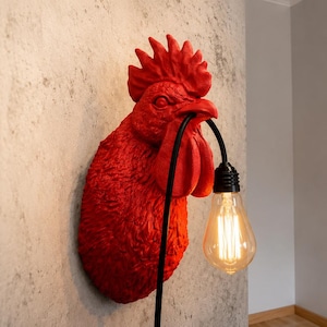 Może przedstawiać: Czerwona lampa ścienna z głową koguta z czarnym przewodem i dużą żarówką w stylu Edisona. Kogut jest zamontowany na teksturowanej szarej ścianie. W tle widoczny jest mały kinkiet ze szklanym kloszem.