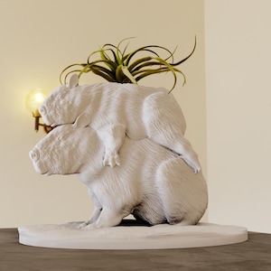 May include: White ceramic sculpture of two capybaras, one on top of the other, with a small green plant growing out of the top capybara's back.