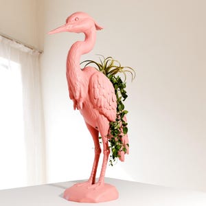 May include: A pink ceramic heron planter with green plants growing out of the top. The heron is standing on a white surface.
