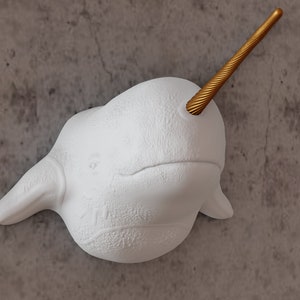 May include: White ceramic narwhal sculpture with a gold-coloured tusk. The sculpture is mounted on a grey textured wall.