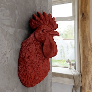 Puede incluir: Una escultura de pared con forma de cabeza de gallo en un acabado rojo mate sólido. La pieza presenta un plumaje detallado y texturizado en el cuello, una cresta prominente en la parte superior y una barbilla distintiva bajo el pico, todo en un solo color.