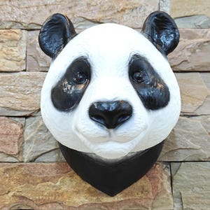 Könnte beinhalten: Pandabären-Kopfskulptur mit schwarzen Ohren, Augenpartien und Nase sowie einem weißen Gesicht. Die Skulptur hat ein schwarzes Halsstück und ist an einer strukturierten, mehrfarbigen Steinmauer befestigt.