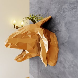 May include: A geometric, golden brown, animal head wall sculpture with a small plant growing out of the top. The sculpture is mounted on a gray wall.