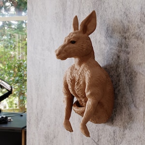 May include: A brown kangaroo wall sculpture. The kangaroo is posed with its arms crossed and is mounted on a textured, light gray wall. The sculpture is made of a light brown material and has detailed features.