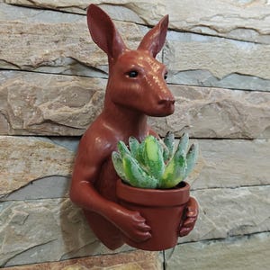 Könnte beinhalten: Ein rotbrauner Wandtopf in Känguruform, der einen kleinen Terrakottatopf mit einer grün-gelben Sukkulente hält. Der Pflanztopf ist an einer steinartigen Wand befestigt.