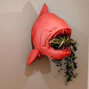 May include: A bright red, 3D printed fish planter with a large open mouth. The fish is mounted on a wall and has a variety of green plants growing out of its mouth.
