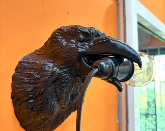 May include: A dark brown raven head wall lamp with a black bulb socket and a clear light bulb. The lamp is mounted on an orange wall. The lamp's cord hangs down. The raven's beak holds the bulb socket.