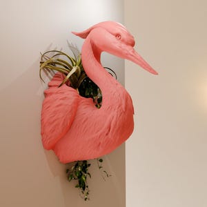 May include: A pink flamingo-shaped planter with a green plant growing inside. The planter is mounted on a white wall.