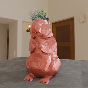 Puede incluir: Un macetero geométrico de color terracota con forma de animal sentado, posiblemente un oso o un perro, con una pequeña planta suculenta que crece en la parte superior.