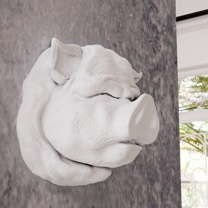 May include: White ceramic pig's head wall decoration. The pig has its eyes closed and its snout is slightly turned to the side. The pig is mounted on a grey wall.