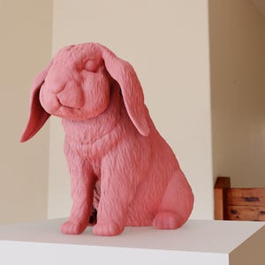 May include: A pink ceramic sculpture of a rabbit sitting with its eyes closed. The sculpture is detailed with fur and has a realistic appearance.