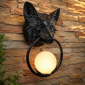 Könnte beinhalten: Schwarze Fuchskopf-Wandleuchte mit einer runden, leuchtenden Glühbirne. Der Fuchskopf ist an einer strukturierten, braunen Steinmauer befestigt. Das Licht wird in einem schwarzen Ring gehalten, was eine einzigartige dekorative Leuchte ergibt.