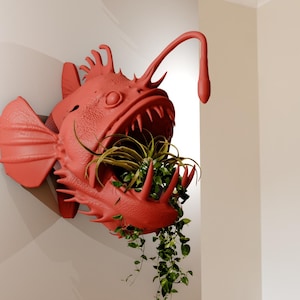 Puede incluir: Un macetero de pared de pez pescador rojo con un apéndice largo, delgado e curvo que se extiende desde la parte superior de su cabeza. La boca del pez pescador está abierta y contiene una variedad de plantas verdes.