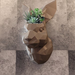 May include: A brown geometric rabbit head planter with a variety of green succulents growing inside. The planter is mounted on a gray tiled wall.