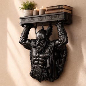 Peut inclure: Une étagère murale noire représentant une sculpture de guerrier barbu avec un casque ailé soutenant l'étagère au-dessus de sa tête. Le bord de l'étagère est orné de symboles runiques sculptés et porte une petite plante en pot et deux livres.