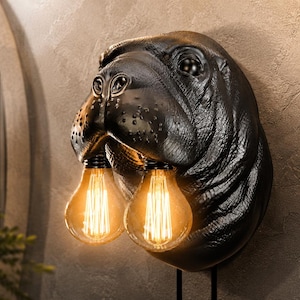 Könnte beinhalten: Eine Wandleuchte in Form eines schwarzen Nilpferdkopfes mit matter, texturierter Oberfläche. Zwei leuchtende, klare Glühbirnen im Edison-Stil hängen am unteren Teil der Schnauze des Nilpferdes und strahlen ein warmes, bernsteinfarbenes Licht aus.