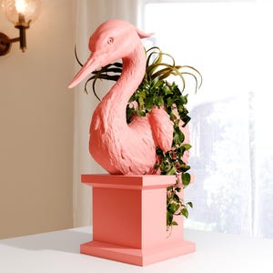 May include: A pink ceramic bird planter with a green plant growing out of it. The planter is on a pink pedestal.