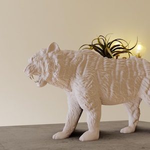 以下が含まれることがあります： 小さな緑色の植物が植えられた白いセラミックの虎のプランター。プランターはリアルな毛皮と唸る表情で詳細に描かれています。