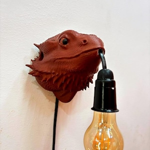 May include: A unique wall-mounted light fixture shaped like a reddish-brown lizard head. The lizard's mouth holds a black light socket with a vintage-style Edison bulb. A black cord extends down from the fixture.