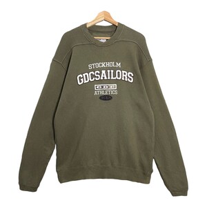 Puede incluir: Sudadera verde oliva con el texto "STOCKHOLM GDCSAILORS ATHLETICS" en blanco y negro. La sudadera está colgada de una percha de madera. El diseño está centrado en el pecho.