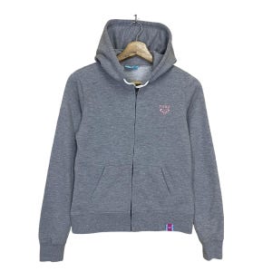 May include: A grey zip-up hoodie with a pink Roxy logo on the front. The hoodie has a hood with a drawstring and a kangaroo pocket.