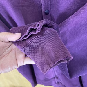May include: Close-up of a purple sweatshirt sleeve with a ribbed cuff. The fabric is a solid, deep purple color. The sleeve is slightly worn, with some fraying at the cuff edge. A dark blue button is visible.