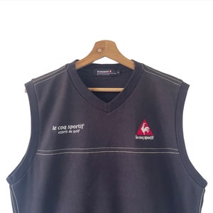 May include: Black sleeveless sweater vest with a v-neck. The vest has a white embroidered logo that says "le coq sportif esprit de golf" on the left side and a red rooster logo with "le coq sportif" text on the right side.