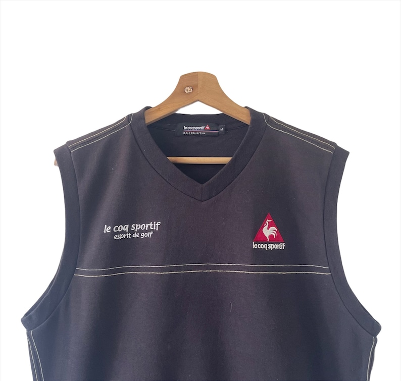 May include: Black sleeveless sweater vest with a v-neck. The vest has a white embroidered logo that says "le coq sportif esprit de golf" on the left side and a red rooster logo with "le coq sportif" text on the right side.