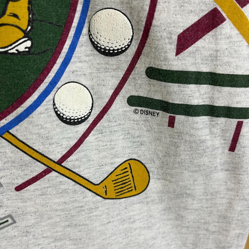 Puede incluir: Primer plano de un gr&aacute;fico gris con tem&aacute;tica de golf. El dise&ntilde;o incluye dos pelotas de golf blancas, un palo de golf amarillo y l&iacute;neas verdes, azules y granates. El texto "&copy; DISNEY" es visible.