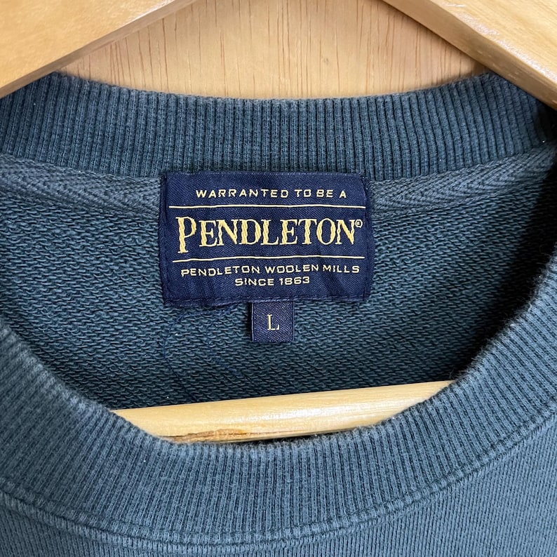 以下が含まれることがあります： 濃いティール色のペンドルトン スウェットシャツのクローズアップ。ネイビーブルーのラベルには「WARRANTED TO BE A PENDLETON&reg; PENDLETON WOOLEN MILLS SINCE 1863」とサイズ「L」が記載されています。スウェットシャツはリブ編みのクルーネックです。
