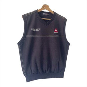 May include: A black sleeveless sweater vest with a v-neck. The vest has a white embroidered line across the chest and the text "le coq sportif esprit de golf" and a red rooster logo with the text "le coq sportif" on the right side.