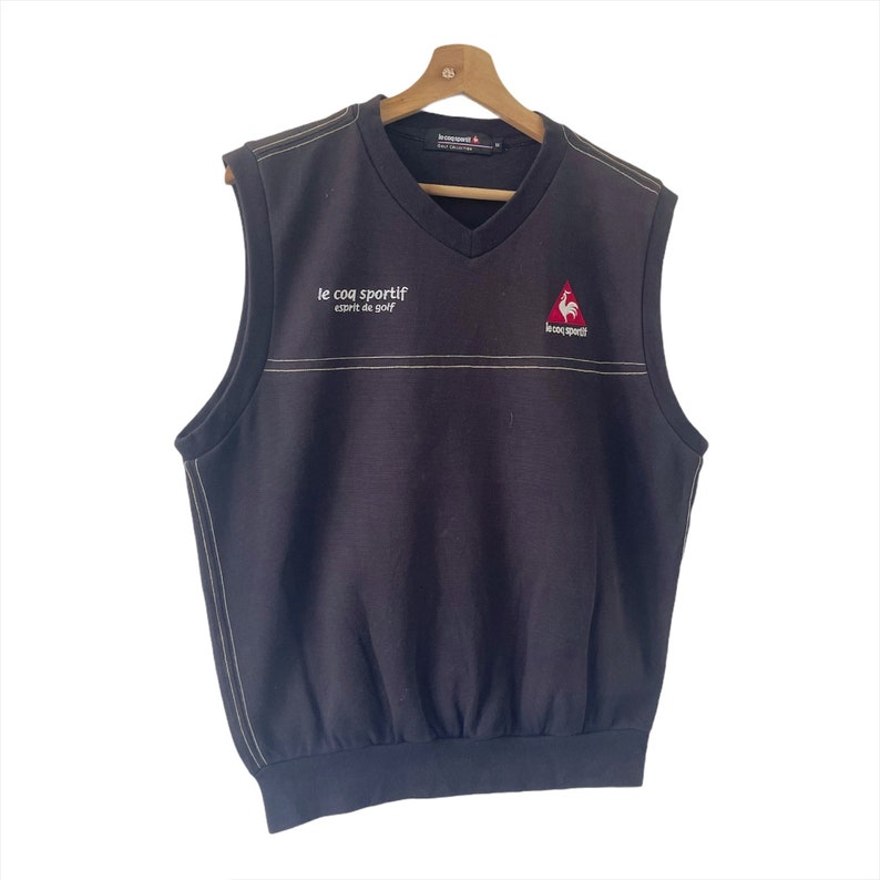 May include: A black sleeveless sweater vest with a v-neck. The vest has a white embroidered line across the chest and the text "le coq sportif esprit de golf" and a red rooster logo with the text "le coq sportif" on the right side.
