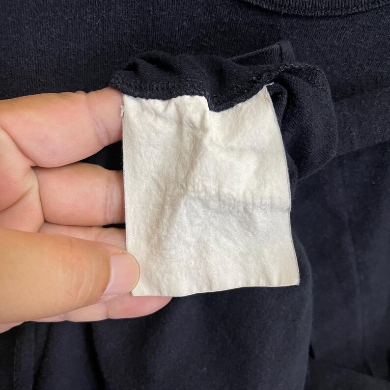 May include: Close-up of a black garment with a white tag attached. The tag is rectangular and appears to be made of a soft, textured material. The garment's fabric is dark and slightly wrinkled.