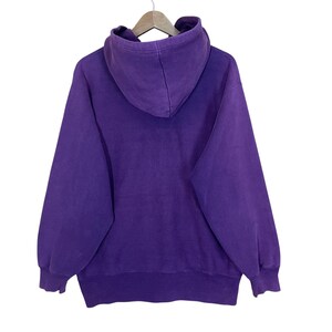 May include: A purple hoodie with a hood and long sleeves, hanging on a wooden hanger. The hoodie is a solid color and appears to be made of a soft material. The back of the hoodie is visible.