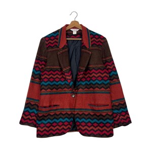 May include: A vintage blazer with a colorful geometric pattern. The jacket features a brown, red, and teal zig-zag design with a single-button closure. The blazer is hanging on a wooden hanger.