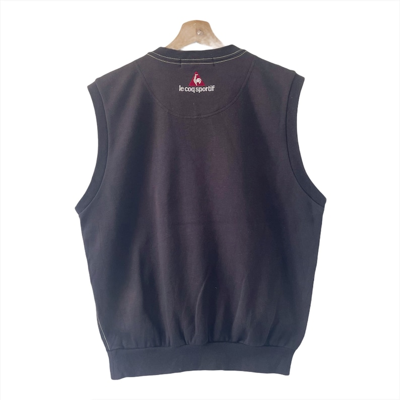 May include: A black sleeveless sweater vest with a white trim around the neck and armholes. The vest has a small red rooster logo with the text "le coq sportif" on the back.