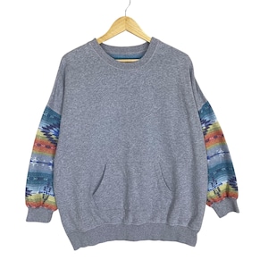 Op de afbeelding: Een grijze sweatshirt met een kangoeroezak en mouwen met patronen. De mouwen hebben een kleurrijk geometrisch ontwerp in tinten blauw, oranje en rood. De sweatshirt hangt aan een houten hanger.
