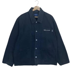 May include: Black jacket with a button-up front, two front pockets, and a collar. The jacket has the text "COMME ÇA DU MODE" printed on the front. The jacket is hanging on a wooden hanger.