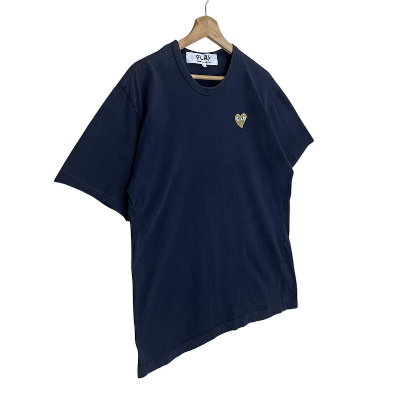 May include: Navy blue t-shirt with a small gold heart-shaped graphic on the chest. The shirt has a crew neck and short sleeves. The word "PLAY" is printed on a small white tag at the neckline.
